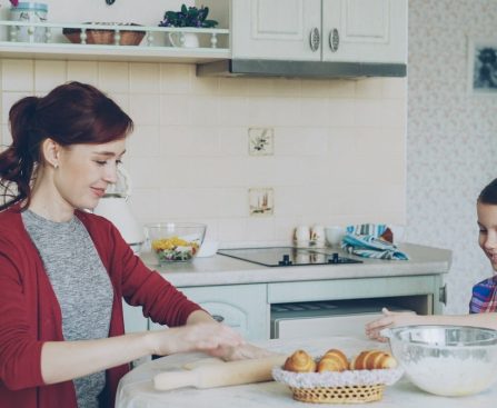 Young mother rolling dough and talking to her little cute daughter while cooking in the kitchen on weekend. Family, food, home and people concept