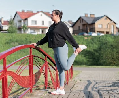 A woman in active wear performs a stretching exercise on a red bridge in a suburban park setting.