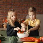 Cheerful girl helping little boy in apron preparing salad while standing in kitchen near table with mix of fresh vegetables behind brick wall