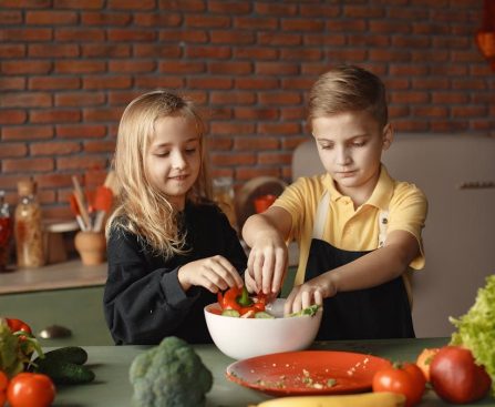 Cheerful girl helping little boy in apron preparing salad while standing in kitchen near table with mix of fresh vegetables behind brick wall